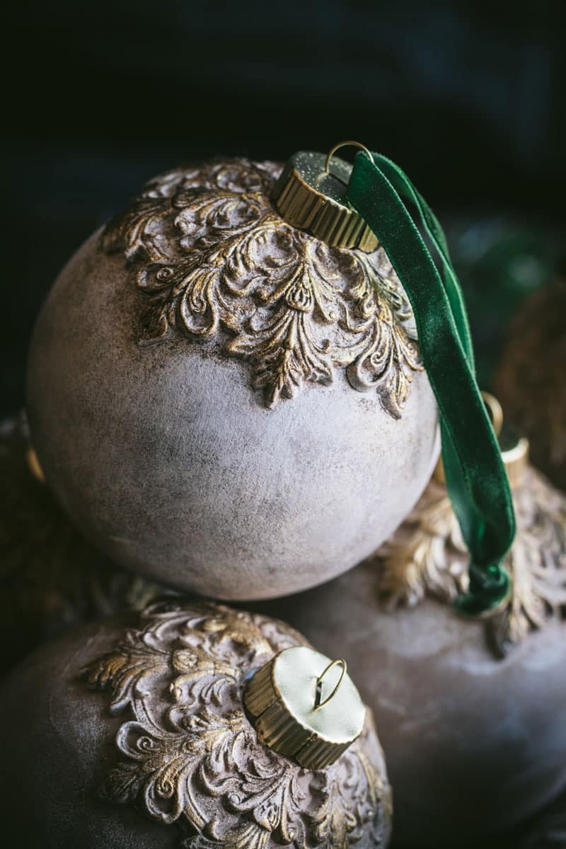 Two ornate Christmas ornaments with gold decorative tops rest together. One ornament has a green velvet ribbon looped through its gold cap, while the other is without a ribbon. The background is dark and softly blurred.