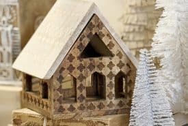 A small decorative wooden house sits on a stack of old books beside two white bottle brush trees and other neutral holiday decor in the background.