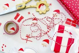Gift wrapping supplies including patterned Santa wrapping paper, red and white string, scissors with floral handles, a red polka dot wrapped gift, tape dispensers, and a wrapped present on a marble surface.