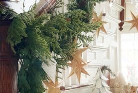 A staircase railing decorated with green garland and wooden star ornaments. At the bottom of the stairs are white holiday decorations, including a tree and gingerbread houses, creating a festive, cozy atmosphere.