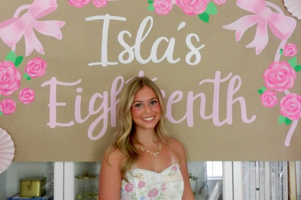 A young woman in a floral dress stands smiling in front of a sign decorated with pink flowers and bows that reads "Isla's Eighteenth." White shelves with books and decor are in the background.