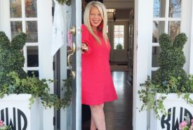 A smiling woman in a bright pink dress stands in an open doorway on a blue porch. The doormat reads "welcome THE WOODS." Two large white planters with leafy greenery and bunny-shaped topiaries flank the door.