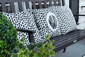 A black metal bench with black and white polka dot pillows, one featuring a monogram. A potted green plant sits in the foreground, and a striped rug is on the gray tile floor. Windows and shutters are in the background.