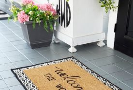 A doormat with the words "welcome THE WOODS" sits on a gray porch near a white planter with greenery and a gray pot of pink flowers. The porch floor has light gray tiles.