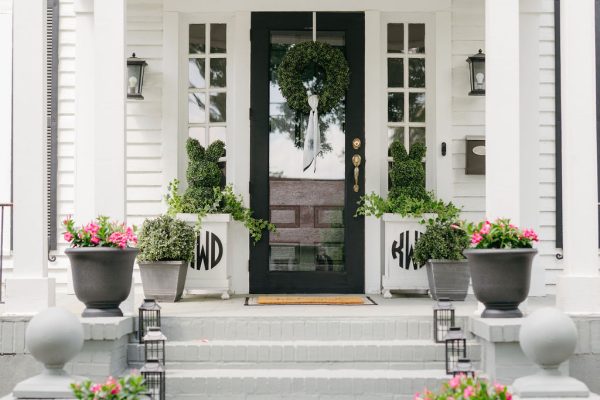 A white house with the address “SEVEN HUNDRED ONE” above the black front door, decorated with green wreaths and potted pink flowers on the steps and porch. Two planters on either side have monogrammed covers.