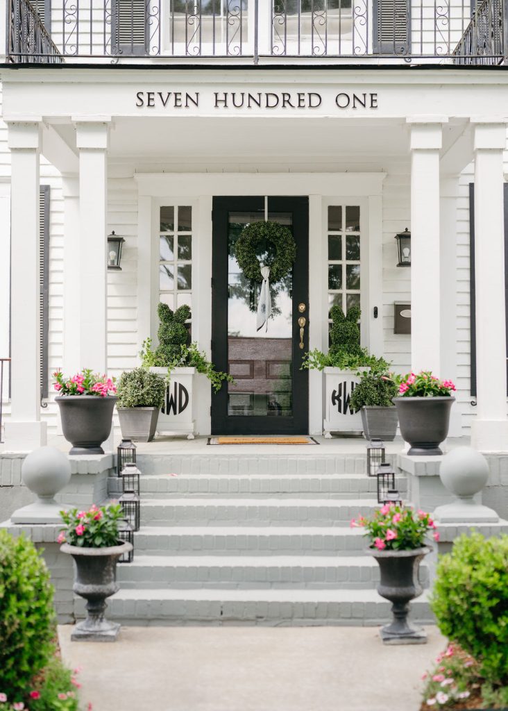 A white house with the address “SEVEN HUNDRED ONE” above the black front door, decorated with green wreaths and potted pink flowers on the steps and porch. Two planters on either side have monogrammed covers.