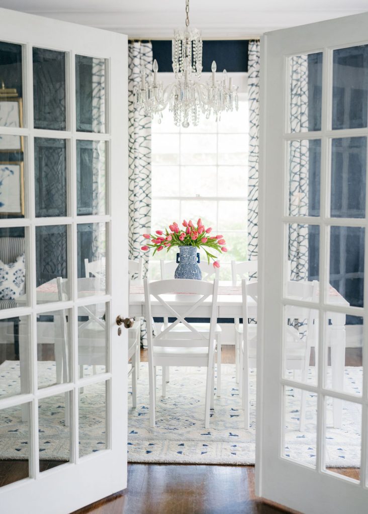 A bright dining room with white French doors, a white table and chairs, a blue vase of pink tulips, a crystal chandelier, patterned curtains, and a large window letting in natural light.