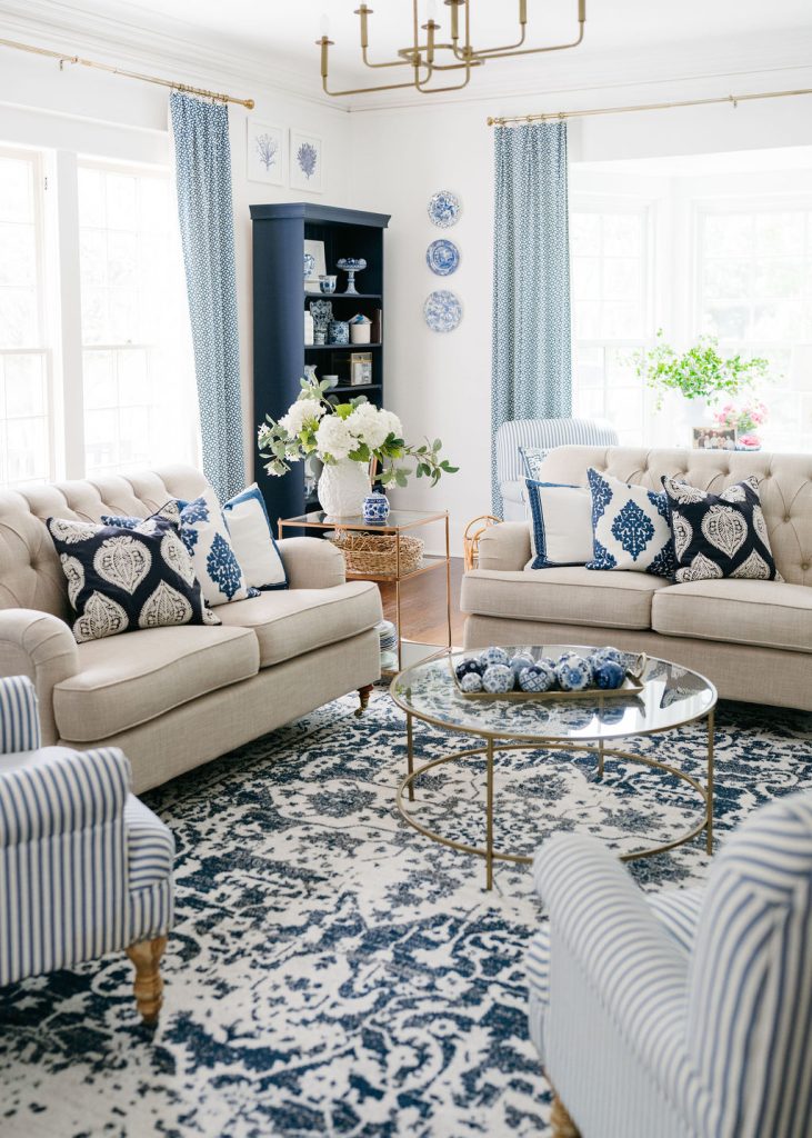 A bright living room with beige sofas, blue and white patterned pillows, striped armchairs, a glass coffee table, a blue patterned rug, blue vases, and large windows with blue curtains letting in natural light.