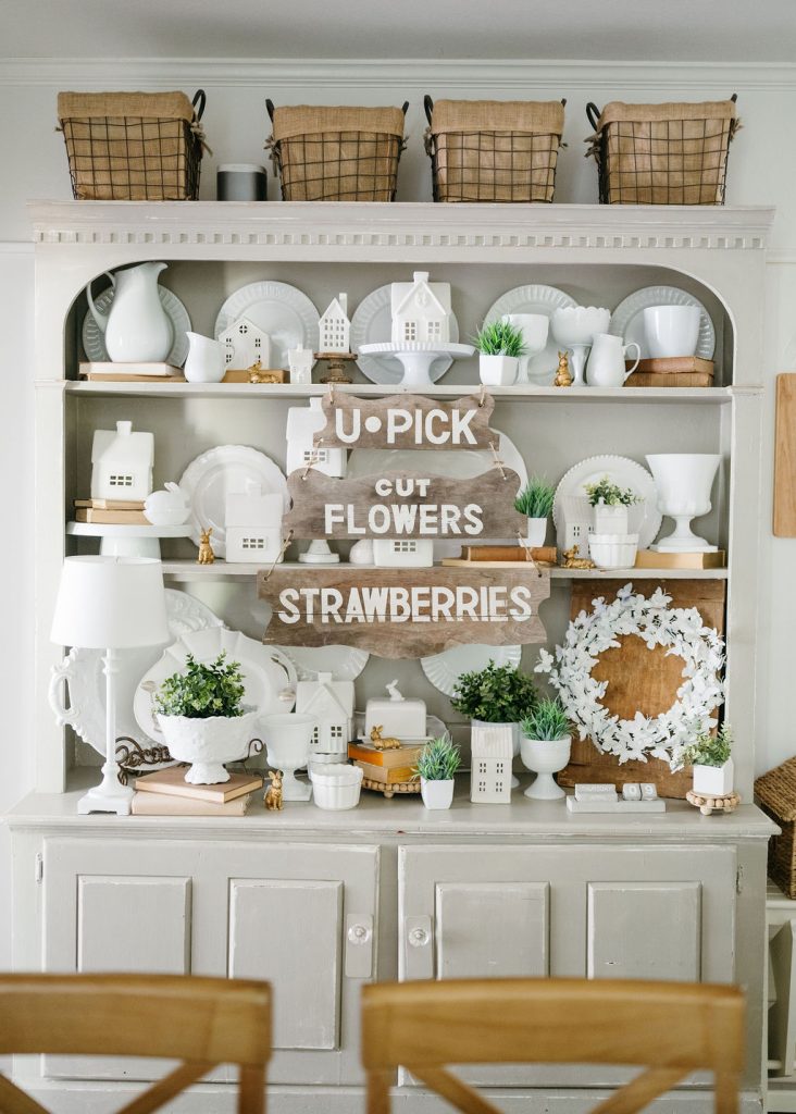 A gray hutch displays white dishes, small potted plants, and farmhouse-style decor. Wicker baskets sit on the top shelf. A rustic sign on the shelves reads "U-PICK CUT FLOWERS STRAWBERRIES.