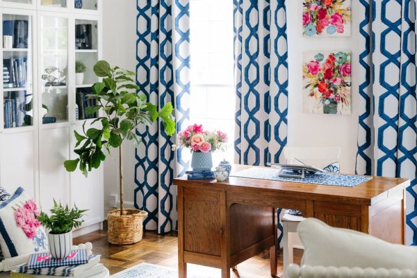 Bright home office with wooden desk, blue and white geometric curtains, floral artwork, a potted plant, bookshelves, and a white armchair. Sunlight streams in, spotlighting blue and white home decor finds and vibrant flowers throughout the room.