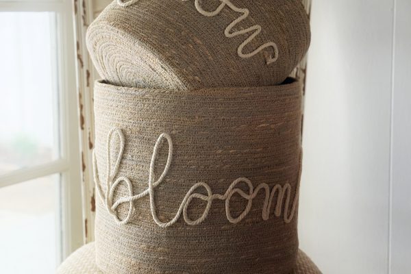 Two woven baskets with white cursive words "grow" and "bloom" stacked on a patterned beige chair, next to a window in a softly lit room.