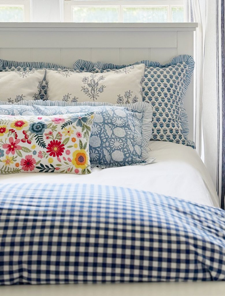 A neatly made bed with white and blue gingham bedding, several decorative pillows in blue and white patterns, and one colorful pillow featuring bright floral designs. Light streams through windows behind the bed.