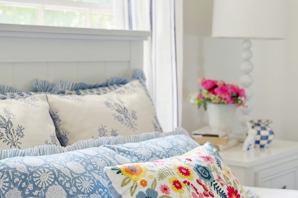 A bright, airy bedroom with a bed featuring blue and white patterned pillows and a colorful floral pillow. A bedside table holds a white lamp, a vase of pink flowers, and a blue-and-white cup. Sunlight filters through the window.