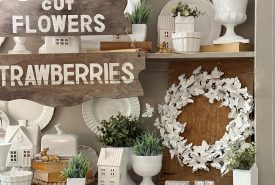 A cabinet displays white dishes, vases, small house figurines, green plants, a white floral wreath, and layered wooden signs reading "U-PICK," "CUT FLOWERS," and "STRAWBERRIES." A small calendar shows "Saturday April 17.