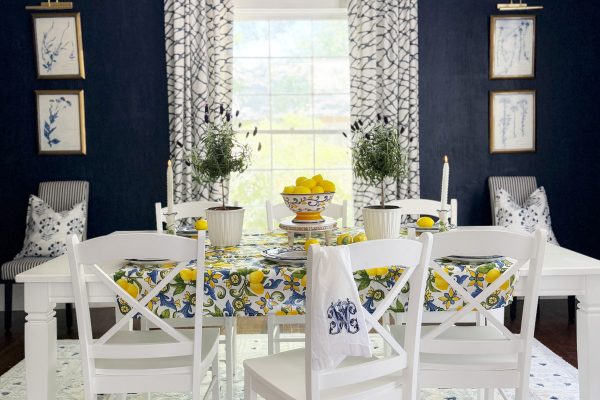 A bright dining room with a white table and chairs, a lemon-themed tablecloth, and a bowl of lemons. Navy blue walls, a chandelier, patterned curtains, and framed art create a fresh, elegant look.
