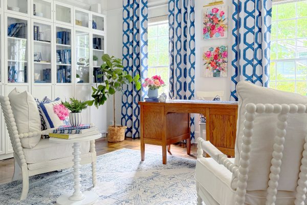 Bright home office with white furniture, a wooden desk, blue and white patterned curtains, a blue and white rug, bookshelves, floral wall art, and potted plants; sunlight streams in through large windows.