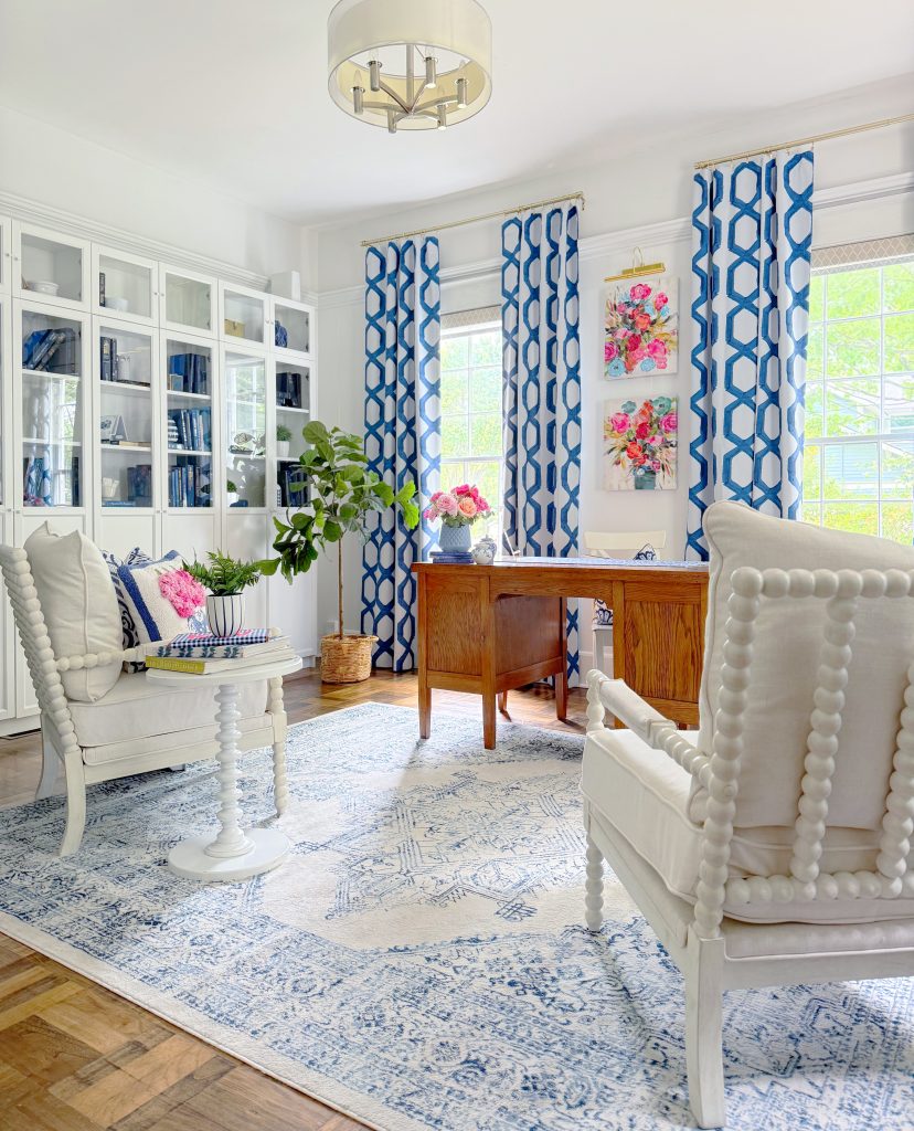 Bright home office with white furniture, a wooden desk, blue and white patterned curtains, a blue and white rug, bookshelves, floral wall art, and potted plants; sunlight streams in through large windows.