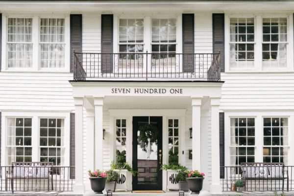 A white two-story house with black shutters, large windows, and a covered front porch. The entrance has potted plants, steps, and a sign above the door that reads "SEVEN HUNDRED ONE." Neatly trimmed shrubs line the walkway.