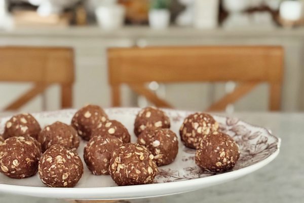 A decorative plate on a stand holds several round chocolate oat energy bites on a marble countertop, with wooden chairs and potted plants in the blurred background.