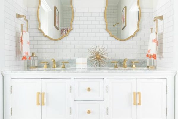 Bright bathroom with two gold-framed mirrors, gold fixtures, white vanity, and three white baskets with pink polka dots underneath. Two pendant lights hang above the sinks, and a gold starburst decor sits between the mirrors.