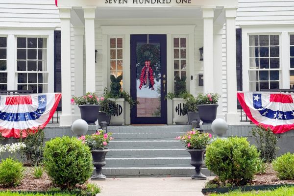 A white house decorated with red, white, and blue patriotic bunting on the porch and balcony, with potted plants by the entrance and manicured bushes lining the walkway.