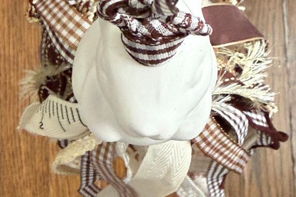 A white ceramic rabbit ornament, decorated with various brown, cream, and plaid fabric ribbons, is hanging against a wooden floor background.