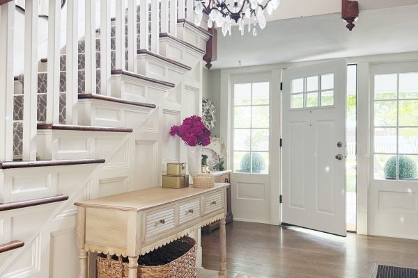 A bright entryway with a staircase, white railings, a light wood console table with a wicker basket, a floral arrangement, and a chandelier. The door and windows let in natural light, illuminating the patterned rug.