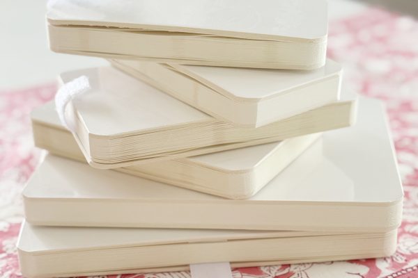 A stack of five white, blank notebooks with rounded edges sits atop a pink and white floral-patterned cloth on a light surface. Two white ribbon bookmarks are visible.