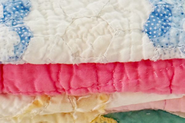 A close-up of a neatly folded stack of colorful handmade quilts, featuring patterns in blue, white, pink, and pastel shades, with visible stitching and soft fabric textures.