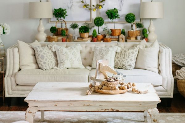 A cozy living room with a white sofa, patterned pillows, and a distressed white coffee table. The wall behind has framed art and a wreath, with potted plants and two lamps on a side table, creating a bright, inviting atmosphere.