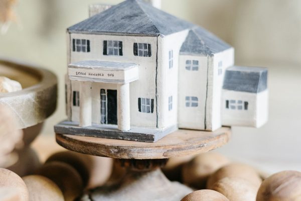 A small, detailed model of a white house with gray roof sits on a wooden stand surrounded by scattered nuts, displayed on a wooden surface indoors.