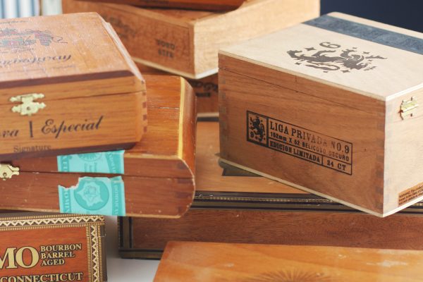 A collection of stacked wooden cigar boxes with various engraved logos and labels, including “Reserva Especial” and “Liga Privada,” arranged on a light surface with a striped chair in the background.