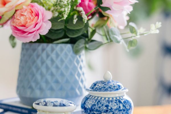 A bouquet of pink and peach flowers in a textured blue vase sits on a wooden table beside two small blue and white porcelain jars with floral patterns.