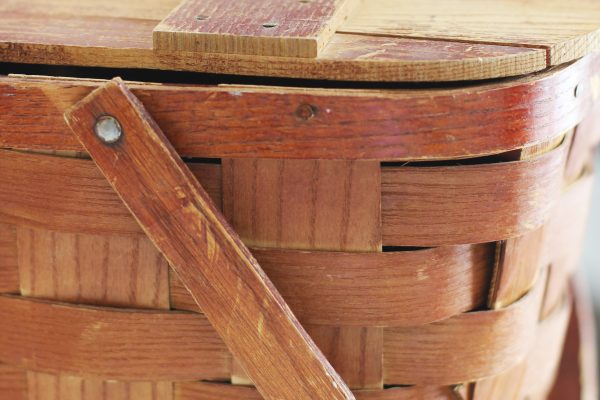 Close-up of a woven wooden picnic basket with a hinged wooden lid and a wooden handle secured by a metal rivet.