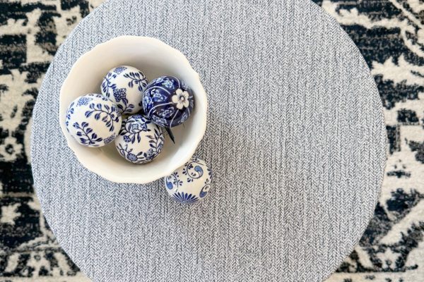 A round, textured gray table with a white bowl holding decorative white and blue patterned balls, placed on a black and white patterned rug.