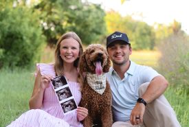 A smiling couple sits on grass with their dog, who wears a "Big Brother" bandana. The woman holds ultrasound photos, suggesting a pregnancy announcement. Trees and greenery are in the background.