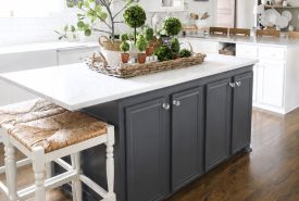 Bright kitchen with a dark gray island, white cabinets, wood floors, wicker stools, and a marble countertop. The island displays a tray with potted plants and greenery under a chandelier. Shelves with dishes are visible in the background.