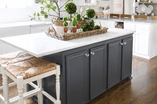 Bright kitchen with a dark gray island, white cabinets, wood floors, wicker stools, and a marble countertop. The island displays a tray with potted plants and greenery under a chandelier. Shelves with dishes are visible in the background.