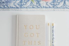 A beige notebook with "YOU GOT THIS" in gold letters lies on a white surface next to two decorative pens. Pink flowers and a blue-patterned cloth are partially visible at the top of the image.
