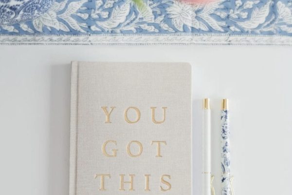 A beige notebook with "YOU GOT THIS" in gold letters lies on a white surface next to two decorative pens. Pink flowers and a blue-patterned cloth are partially visible at the top of the image.