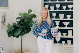 A woman in a blue patterned top and white pants stands smiling in a room, holding two blue and white jars. Behind her is a built-in shelf with white pottery and a green potted plant nearby.