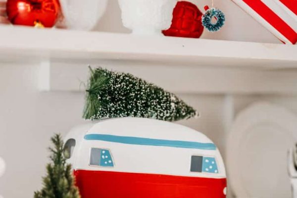 Camper-shaped plates with a toy tree on top sit on a shelf with holiday decor, including a striped gift box, red ornaments, miniature trees, and white dishware.