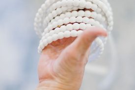 A hand wearing a chunky gold bracelet holds several strands of round, white beads against a soft, blurred background.