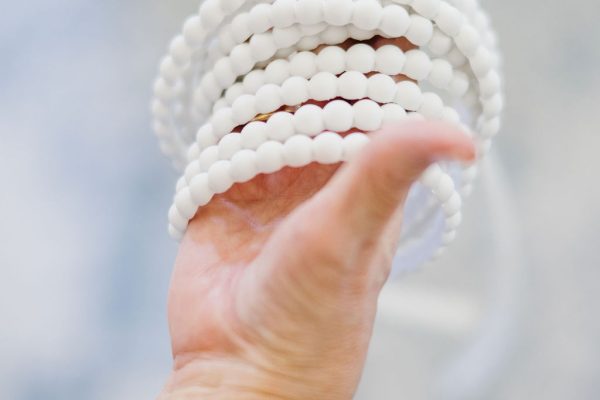 A hand wearing a chunky gold bracelet holds several strands of round, white beads against a soft, blurred background.