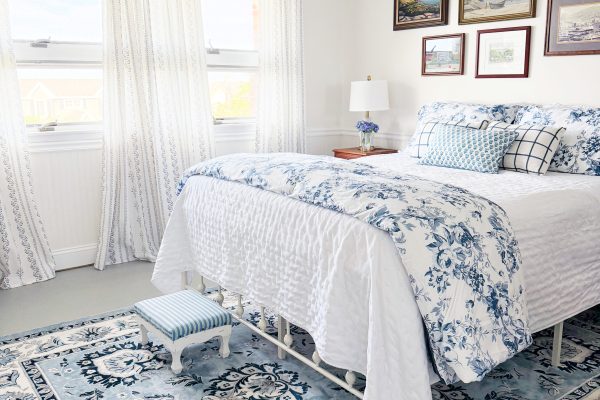 Bright bedroom with a blue and white floral theme, cozy bed, patterned rug, small striped footstool, light curtains, and framed art on the wall. Sunlight fills the room through two large windows.