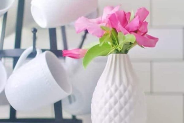 A small white textured vase with pink flowers sits on a countertop. Behind it, white mugs hang on a black metal rack in front of a white subway tile backsplash.