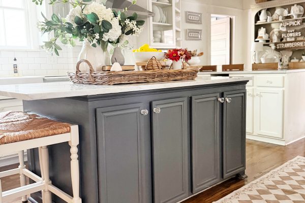 Bright kitchen with a gray island, wicker stools, a chandelier, and a decorative rug. White cabinets, flowers, and baskets add a cozy, farmhouse feel. Natural light streams in from the window.
