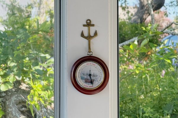 A brass anchor decor is mounted above a round barometer on a white wall between two windows, with green trees and plants visible outside.