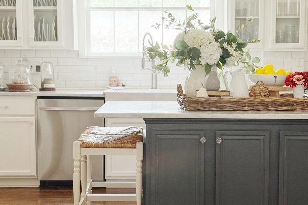 Bright kitchen with white cabinets, a gray island, wicker stools, wood floors, a window with a bamboo shade, a chandelier, fresh flowers in a vase, and lemons in a bowl on the counter.