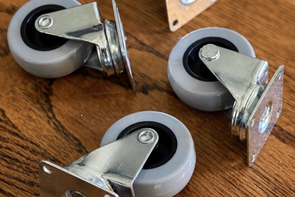 Five gray swivel caster wheels with metal brackets are arranged on a wooden floor.